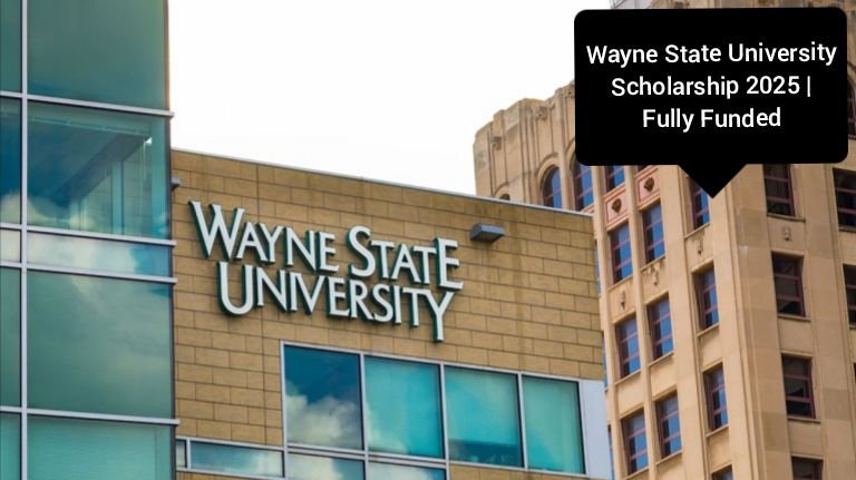 A modern building with large glass windows reflecting the sky and nearby structures. The facade prominently displays the ‘Wayne State University’ signage. Overlay text in the upper right corner reads ‘Wayne State University Scholarship 2025 | Fully Funded,’